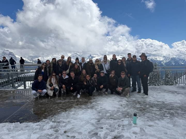 Large tour group poses on a snowy viewing deck with alpine peaks behind.