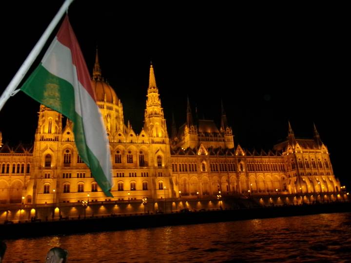 Nighttime view of the illuminated Hungarian Parliament Building with a Hungarian flag in the foreground.