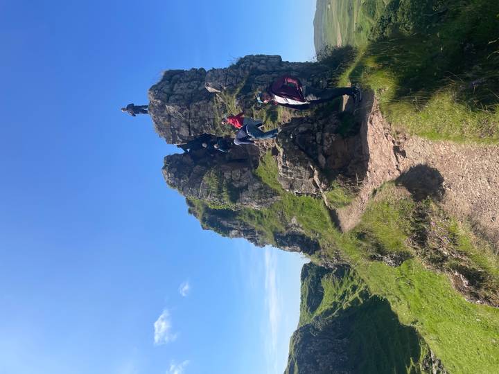 Travellers climb a narrow rocky trail up a craggy green outcrop beneath a bright blue sky.