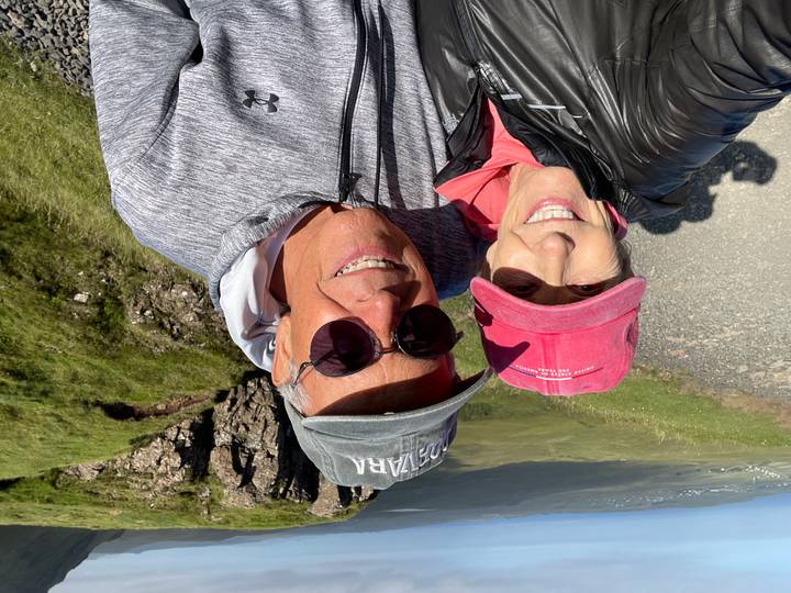 Smiling couple takes a sunny selfie on a scenic Highland roadside overlooking green cliffs.