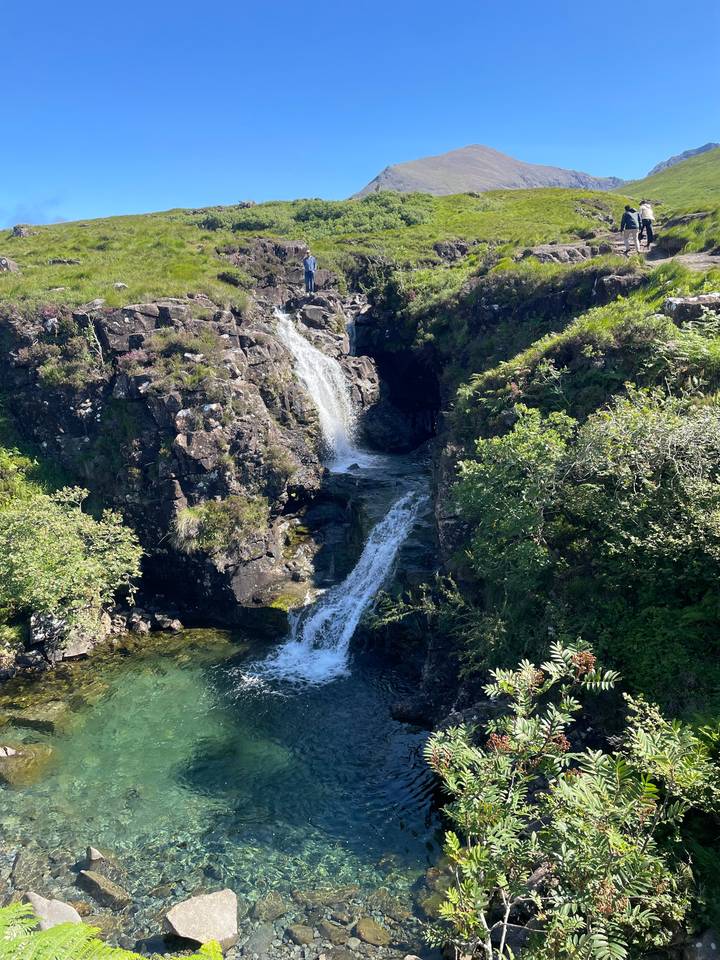 Small waterfall cascades over dark rocks into a clear pool amid lush greenery.