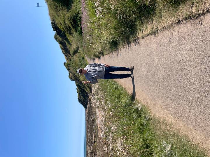 Solo walker on a paved coastal trail bordered by wildflowers and grassy slopes.