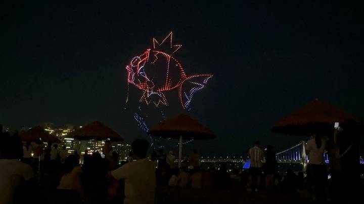 Night beach crowd watching a colourful drone light show forming a fish above the city skyline and bridge.