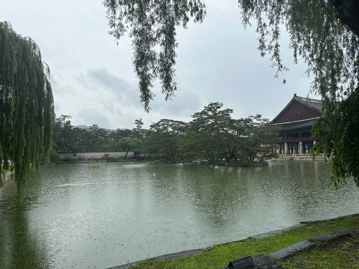 Serene palace pavilion and trees reflected on a rain-dappled pond under overcast sky.