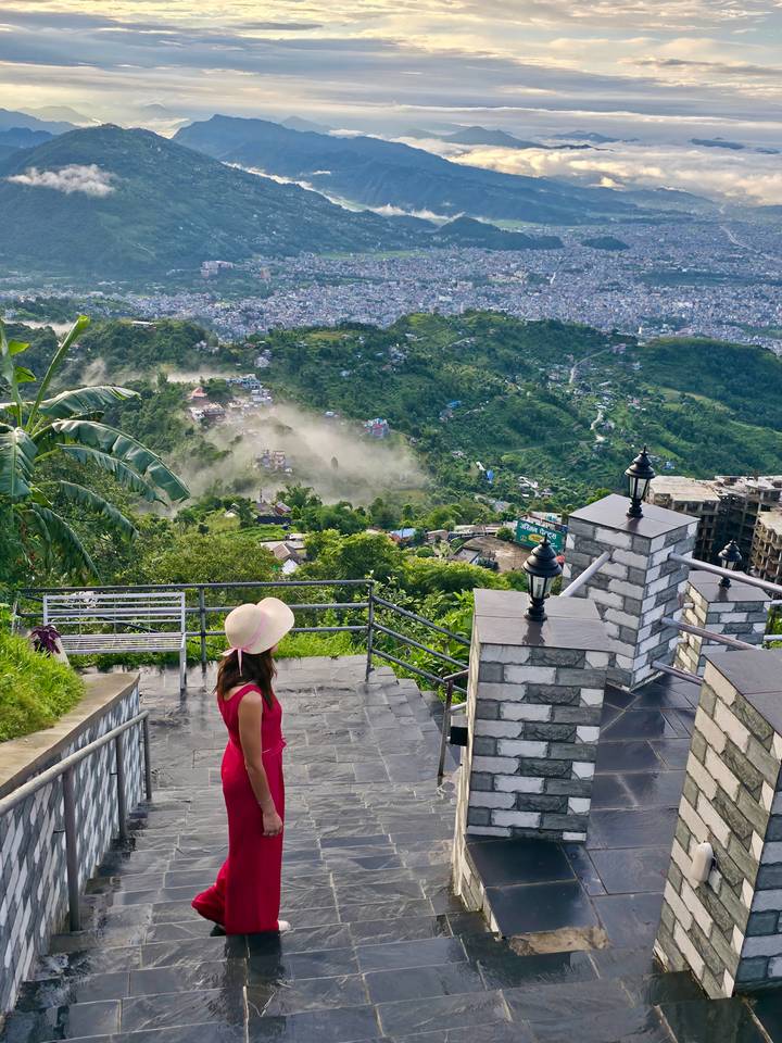 Traveller in pink dress and sunhat admiring lush green valley and distant city from a hilltop viewpoint.