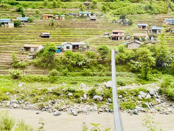Terraced green hills dotted with small houses above a rocky river in rural Nepal.