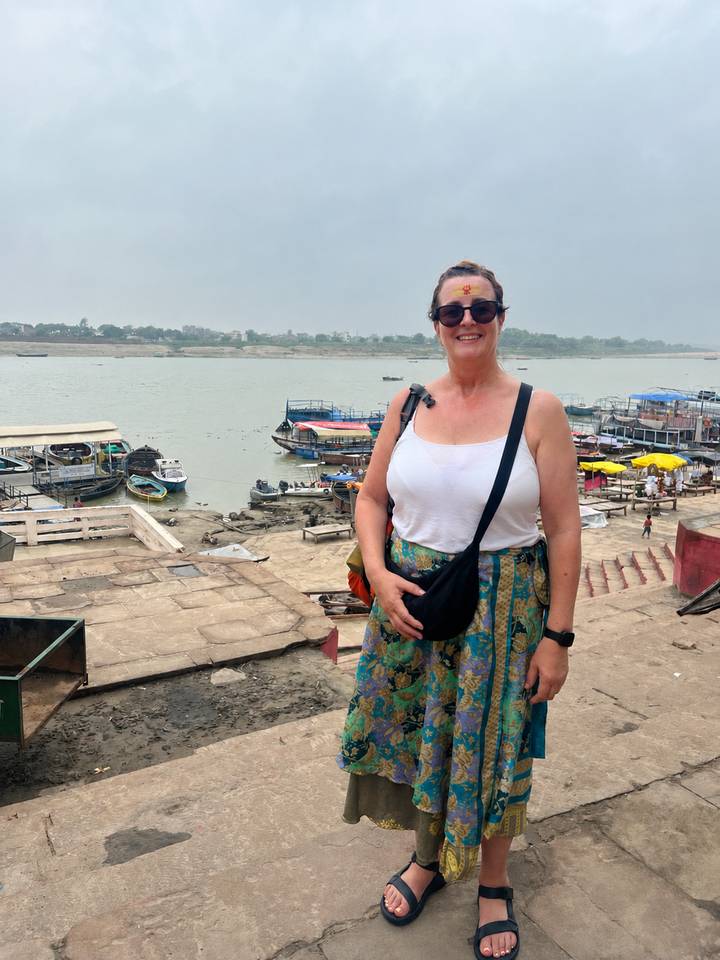A woman with tilak stands on Varanasi ghat steps with boats moored along the Ganges.