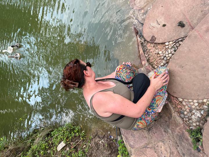 Woman kneels on stones beside a pond reaching toward the water where ducks swim.