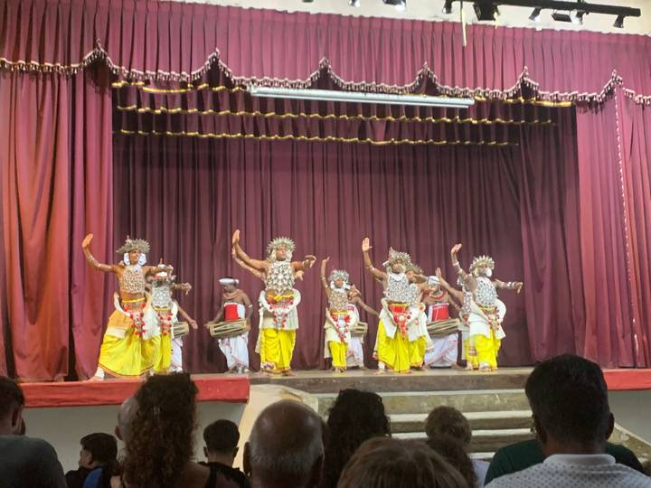 Traditional Sri Lankan dancers in ornate costumes performing on a theatre stage with drummers behind.