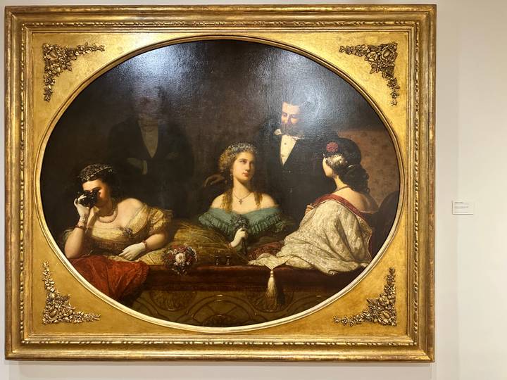 Framed 19th-century salon scene painting displayed on a museum wall under gallery lighting.