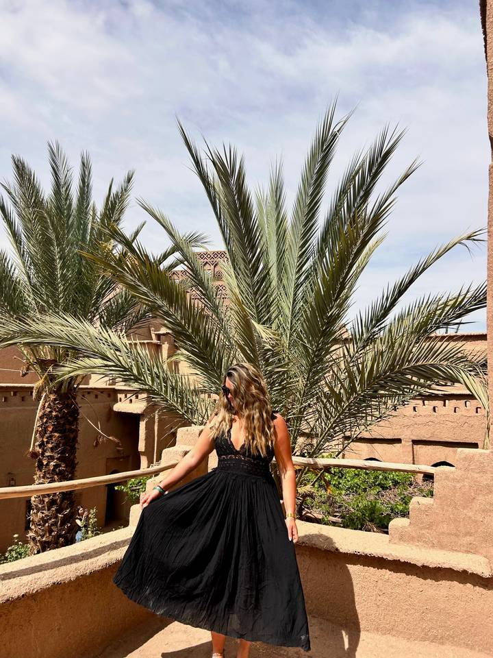Traveler posing by palm trees inside an adobe kasbah courtyard under bright sky.