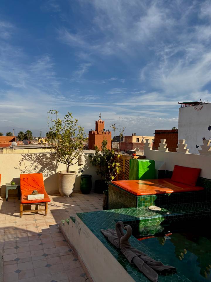 Colorful rooftop terrace in Marrakesh with orange loungers and distant Atlas Mountains.