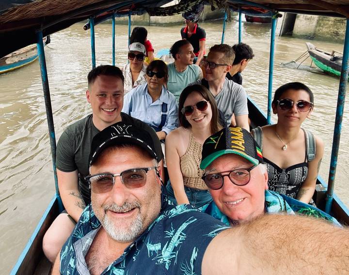 Group selfie of travelers in a narrow boat on muddy Mekong canal wearing casual clothing.