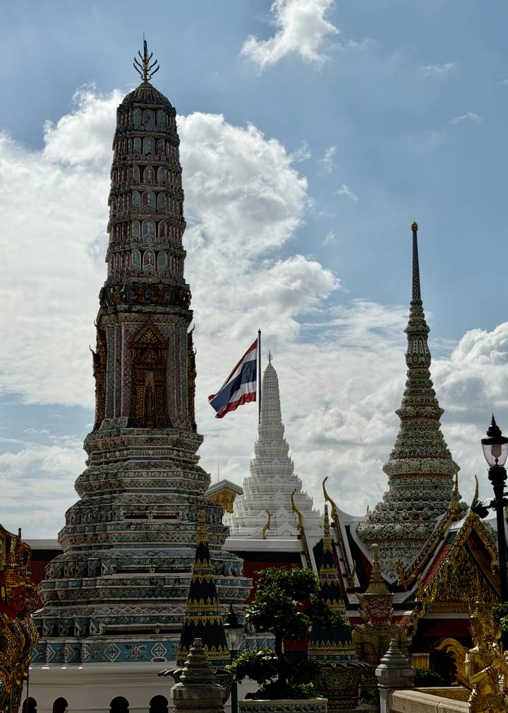 Decorative spires of Wat Arun with a Thai flag fluttering against a blue sky.