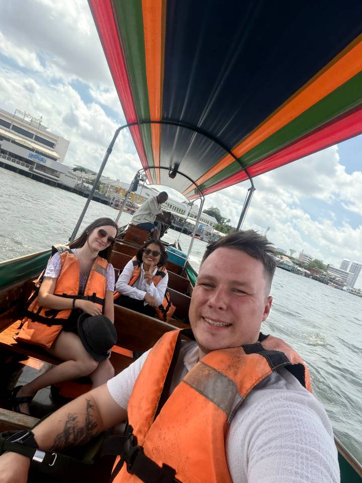 Travellers wearing life jackets smile on a colourful long-tail boat cruising a busy river.