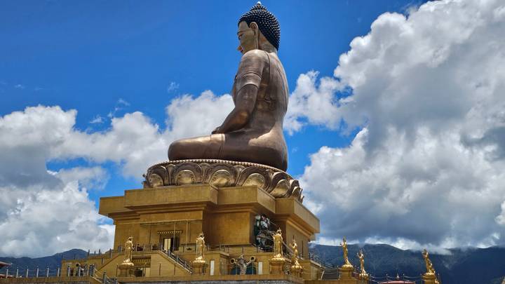 Massive seated bronze Buddha statue atop an ornate golden pedestal set against a bright blue sky with billowing clouds and forested mountains in the background.