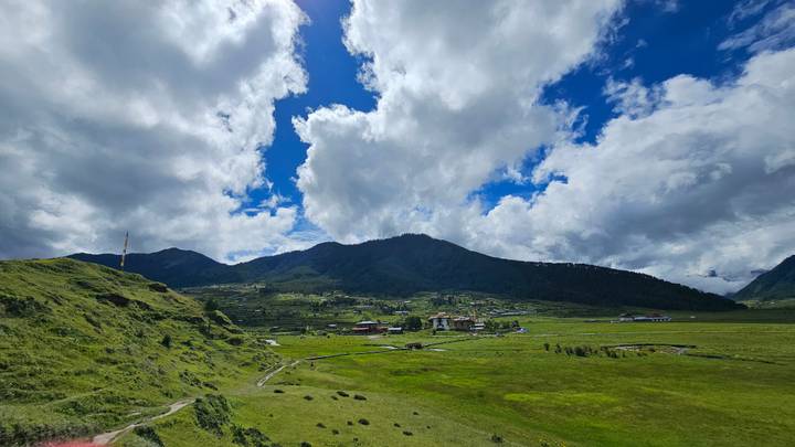 Wide Bhutanese valley with lush green fields, scattered farmhouses and surrounding forested mountains beneath dramatic cloud formations.