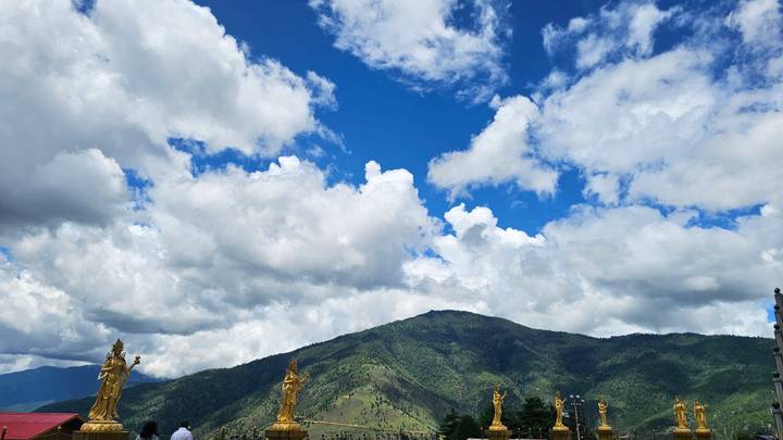 Wide mountain landscape under bright blue sky with dramatic clouds and a row of golden Buddhist statues in the foreground