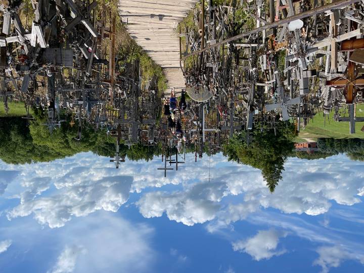 Pathway densely lined with thousands of wooden crosses against a vivid blue cloud-dotted sky
