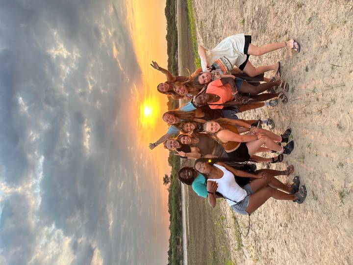 Large group of friends celebrate with arms raised in front of a colorful sunset over open land