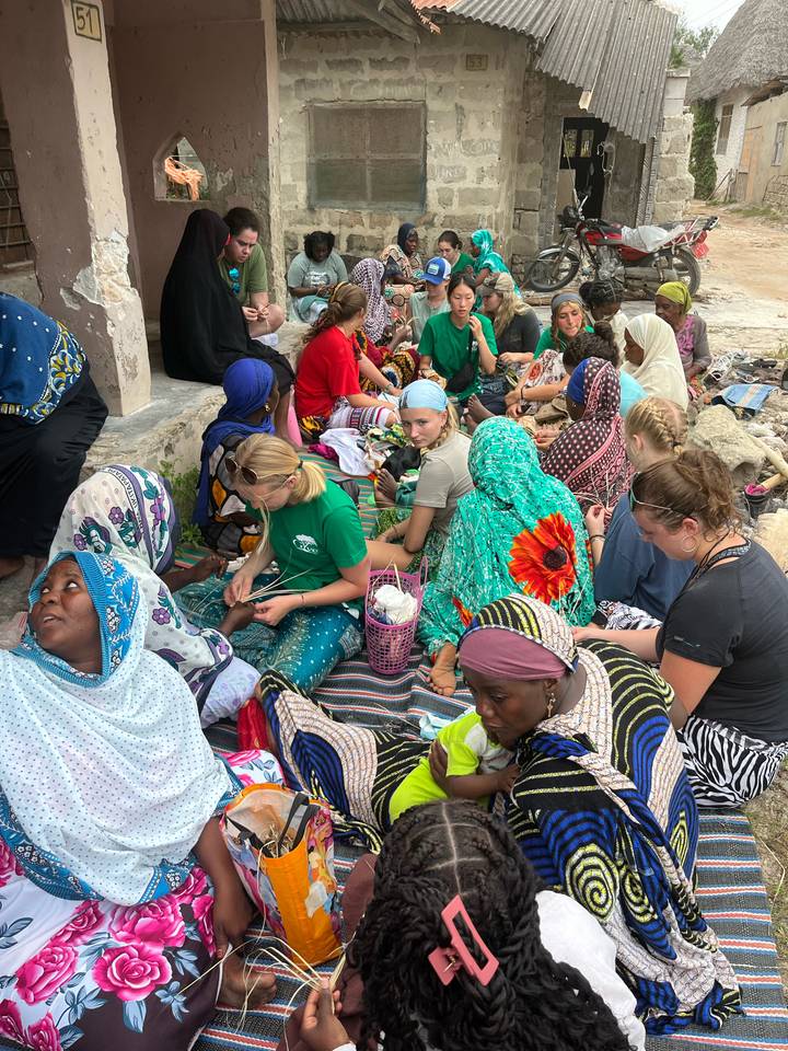 Group of travellers and local women sit together on mats crafting and chatting