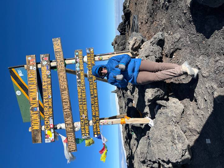 Climber in blue jacket gives thumbs-up beneath the iconic Uhuru Peak sign on snowy Mount Kilimanjaro summit