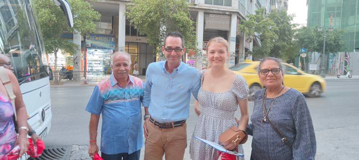 Travellers stand together on a busy city street beside their tour bus, taxis and storefronts behind them.