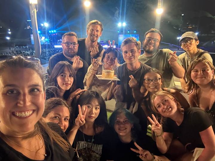 Large group selfie of smiling travelers holding a small cake at an outdoor night event.