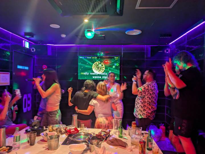 Vibrant karaoke room with travelers singing, dancing and clapping under neon lights.