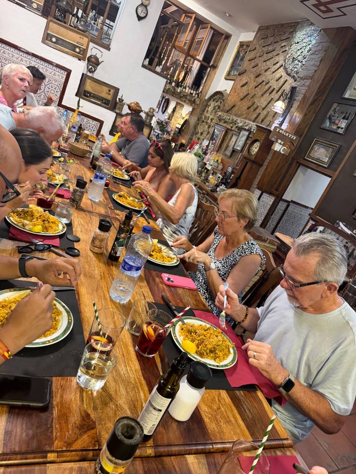 Travelers seated around a wooden table enjoying plates of paella inside a rustic restaurant.