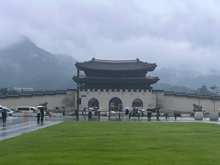 Gwanghwamun Gate stands against misty mountains and rain as visitors with umbrellas cross the plaza.