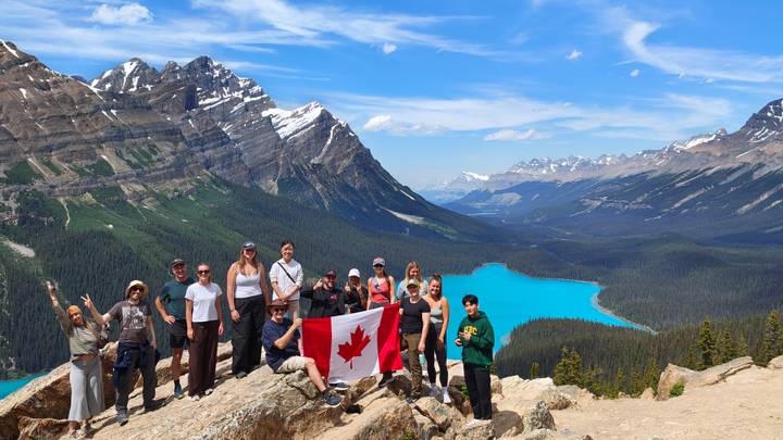 A hiking group waves a Canadian flag atop a rocky viewpoint overlooking turquoise Peyto Lake and the surrounding Rocky Mountains.
