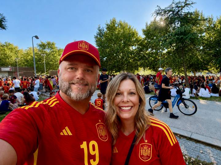 Two enthusiastic football fans in Spain jerseys smile amid a gathering of supporters in a tree-lined plaza.