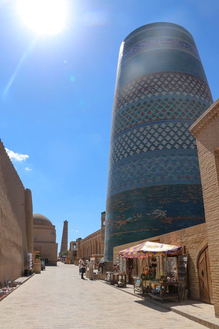 Tall, elaborately tiled minaret rising into a clear blue sky between adobe walls.