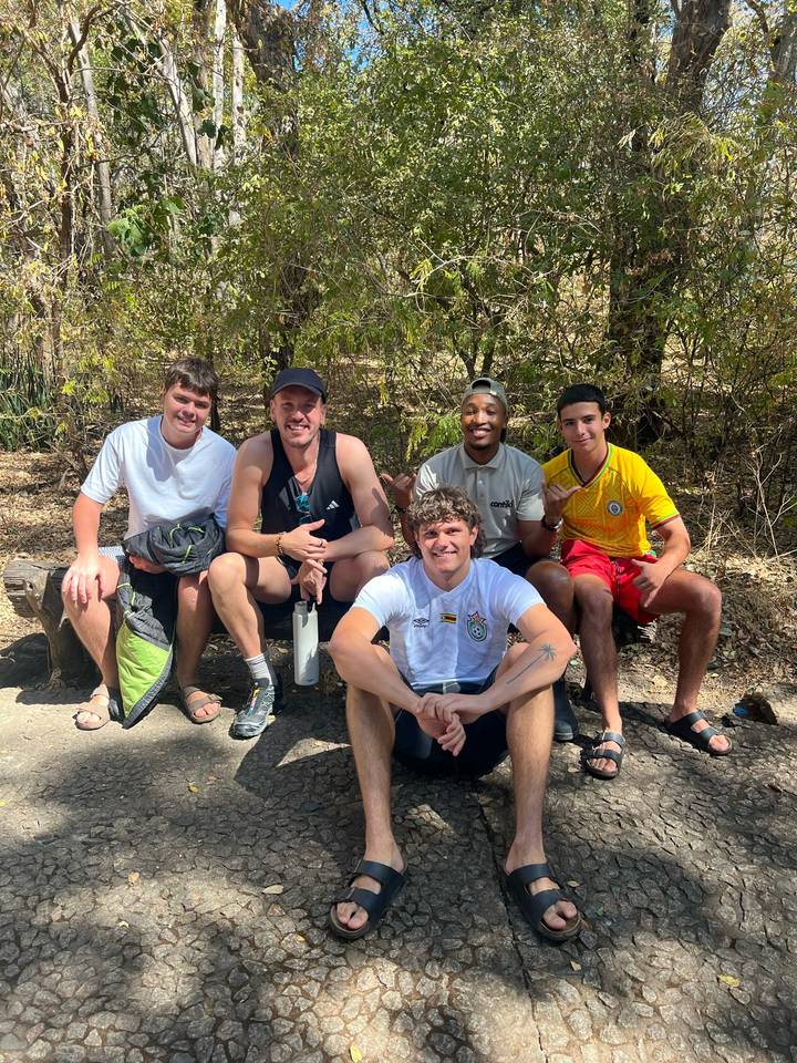 Five young travellers relaxing on a shaded bench in a dry bushveld setting.