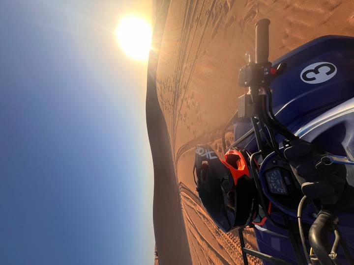 View from an ATV handlebar as it rides across reddish desert sand toward the low sun.