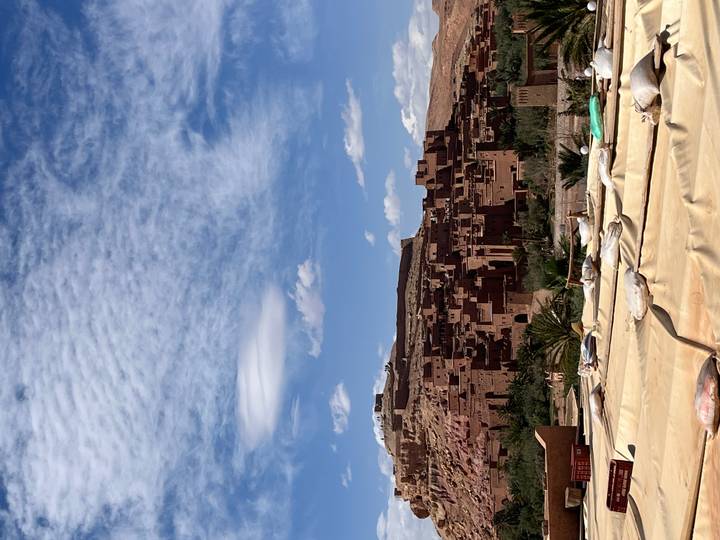 Mud-brick kasbah structures of Ait Benhaddou sprawl beneath a vivid blue sky with dramatic clouds.