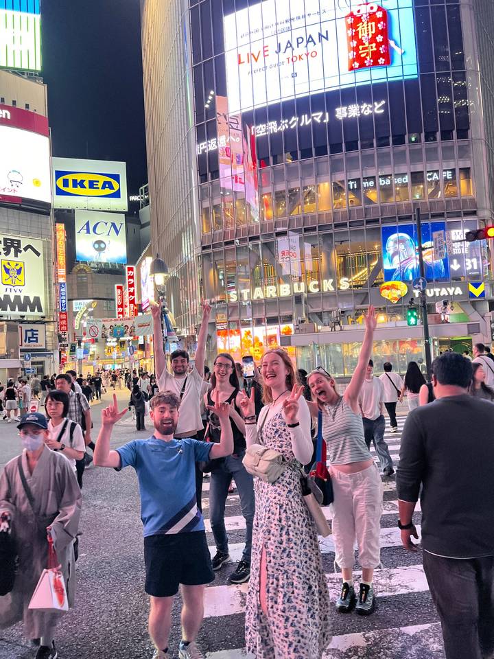 Happy travelers posing with raised arms at Shibuya crossing, neon lights and crowds behind.