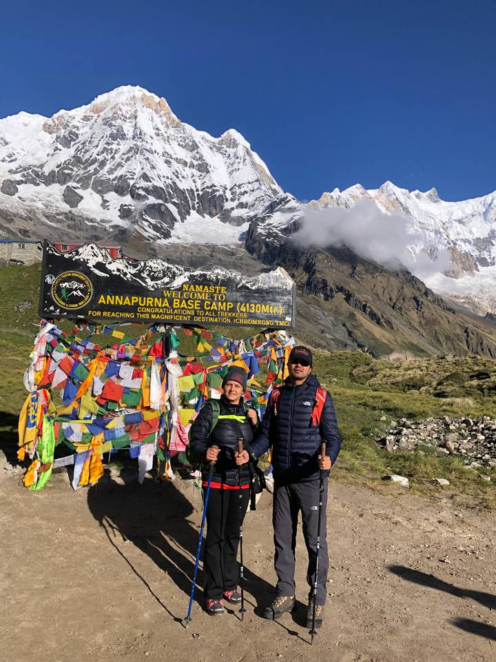Two trekkers posing by the colourful prayer-flag-covered Annapurna Base Camp welcome sign with snowy peaks behind.