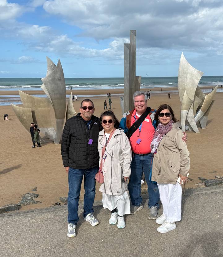 Four smiling travellers pose in front of the stainless-steel ‘Les Braves’ sculpture on a historic beach.