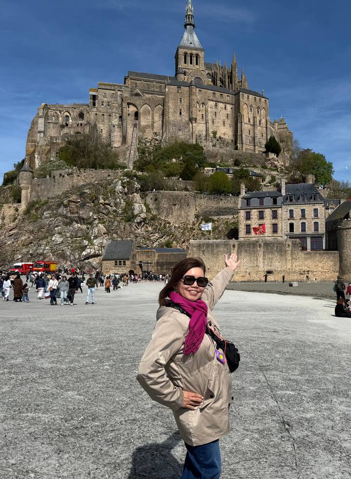Traveller joyfully gestures toward the towering abbey of Mont Saint-Michel with crowds around.