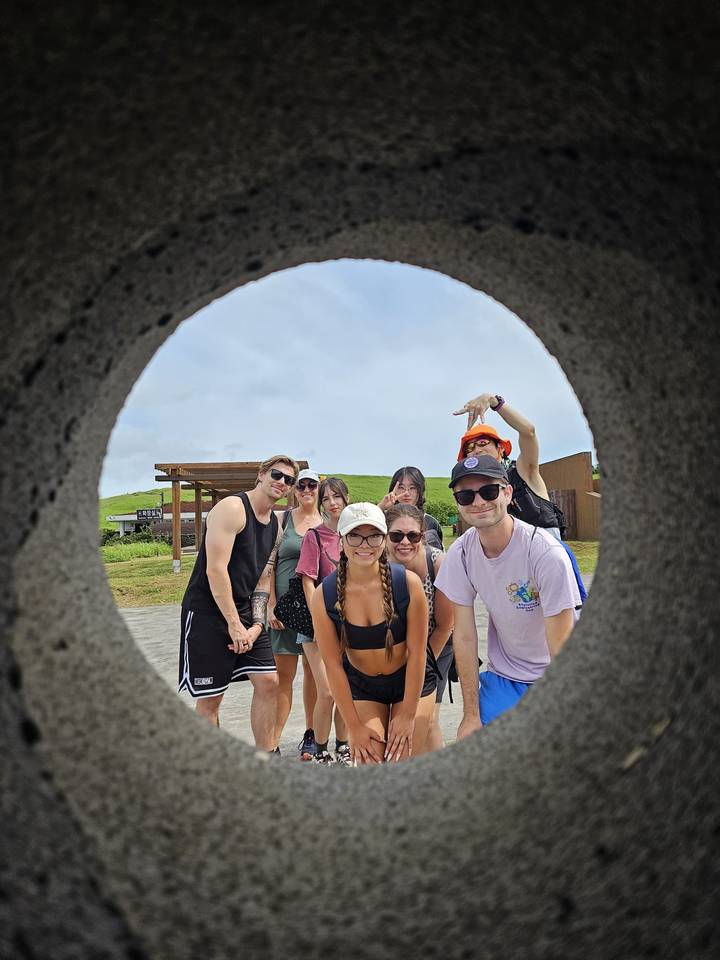 Seven young travelers pose playfully for a group photo through a circular stone frame in a grassy park.