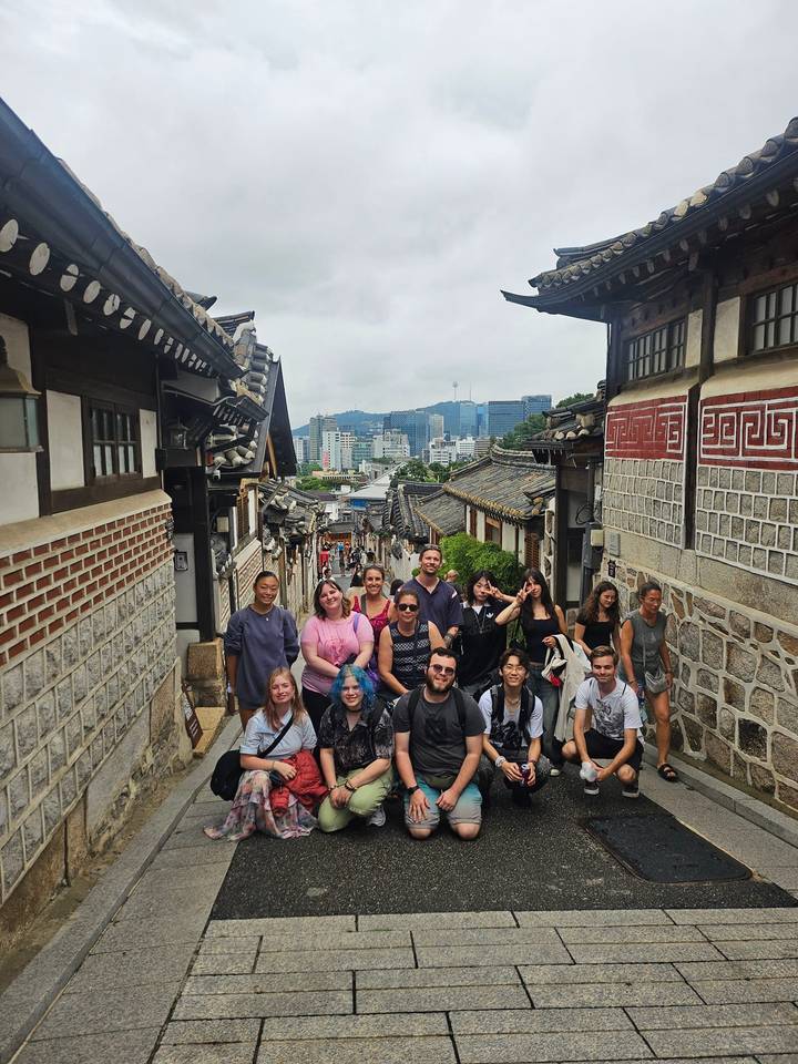 Large tour group gathers on a picturesque stone alley lined with traditional Korean hanok houses overlooking Seoul skyline.