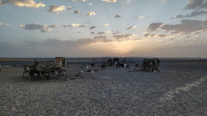 Sparse desert nomad setup with goat pens and wooden shelters silhouetted against a dramatic sunset sky.