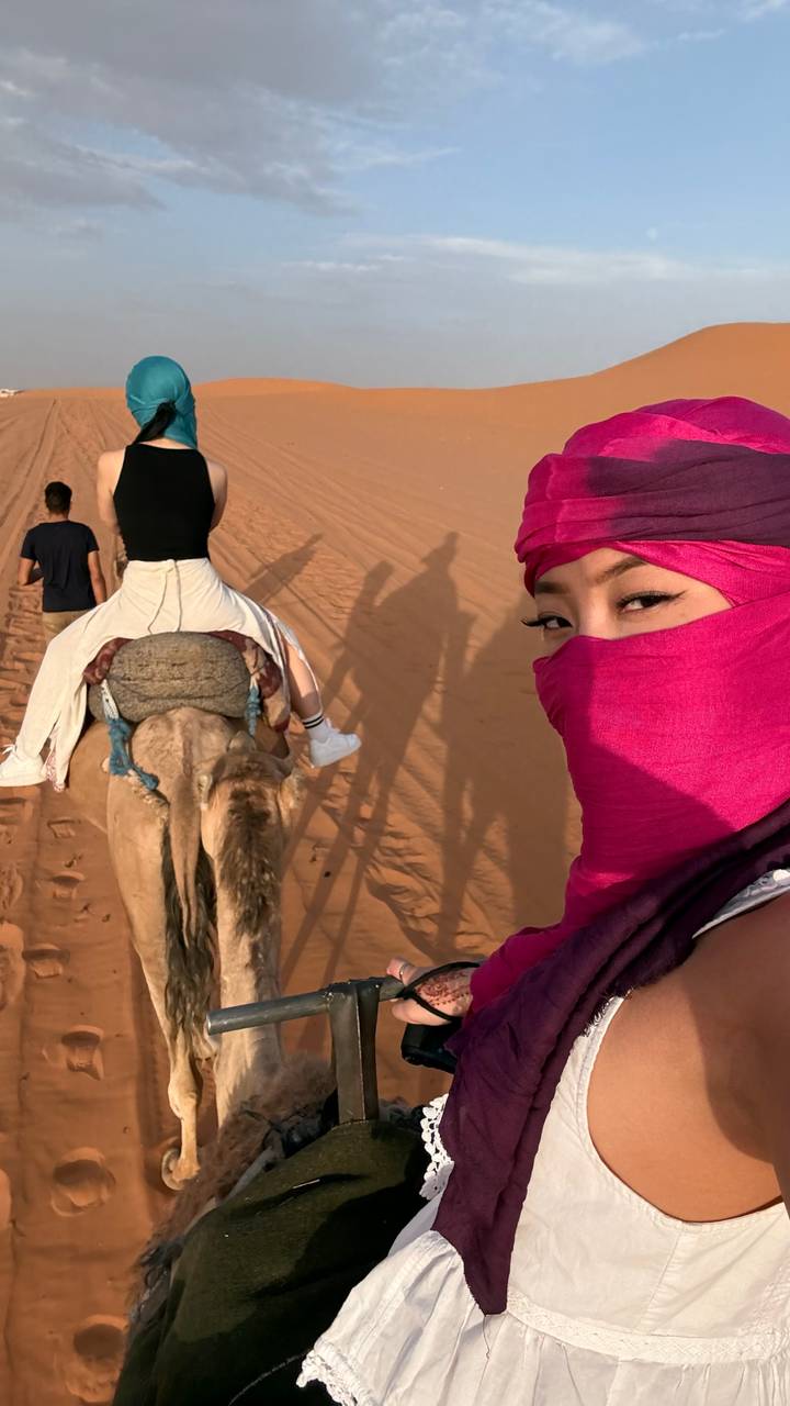 Selfie of traveler in bright desert scarf riding a camel across red sand dunes with companion ahead.