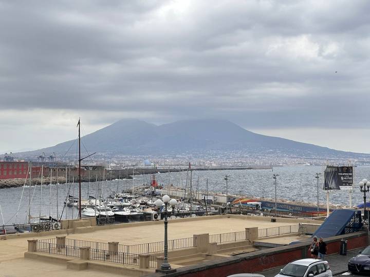 View over a marina and breakwater toward the brooding silhouette of Mount Vesuvius across Naples Bay under gray skies.