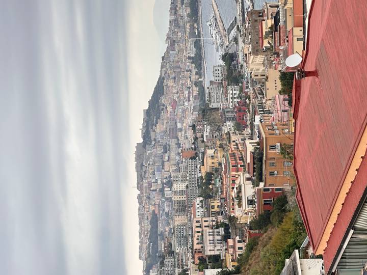 Colorful hillside buildings cascade toward the bay in Naples beneath a cloudy sky.