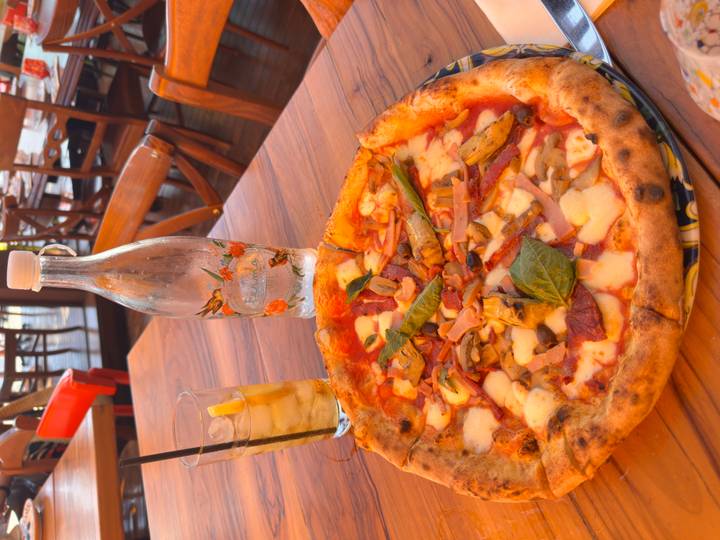Freshly baked Neapolitan pizza topped with vegetables and mozzarella sits on a wooden table beside iced tea and a glass bottle.