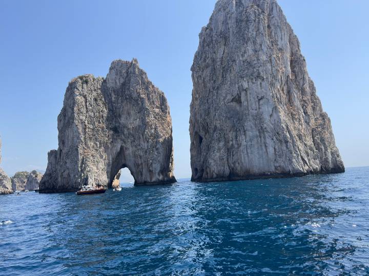 The towering limestone stacks of the Faraglioni rise from deep blue Mediterranean waters seen from a boat.