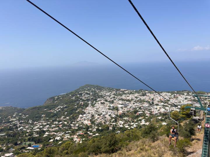 Chairlift cables cut across a high aerial view of Capri island and the endless sea.